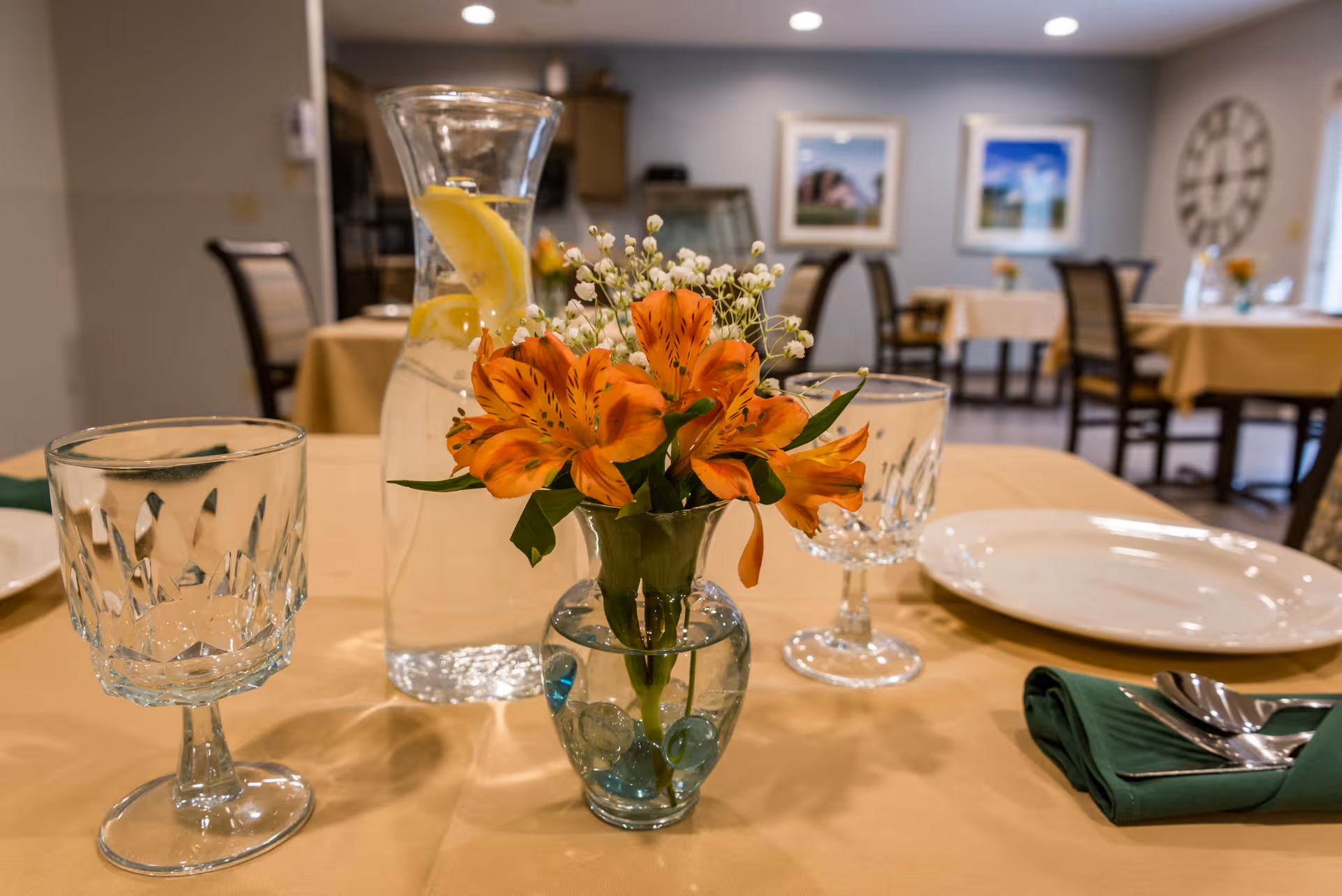 Dining room table set with a small vase of orange flowers, glassware, a pitcher of lemon water, plate, and napkin.