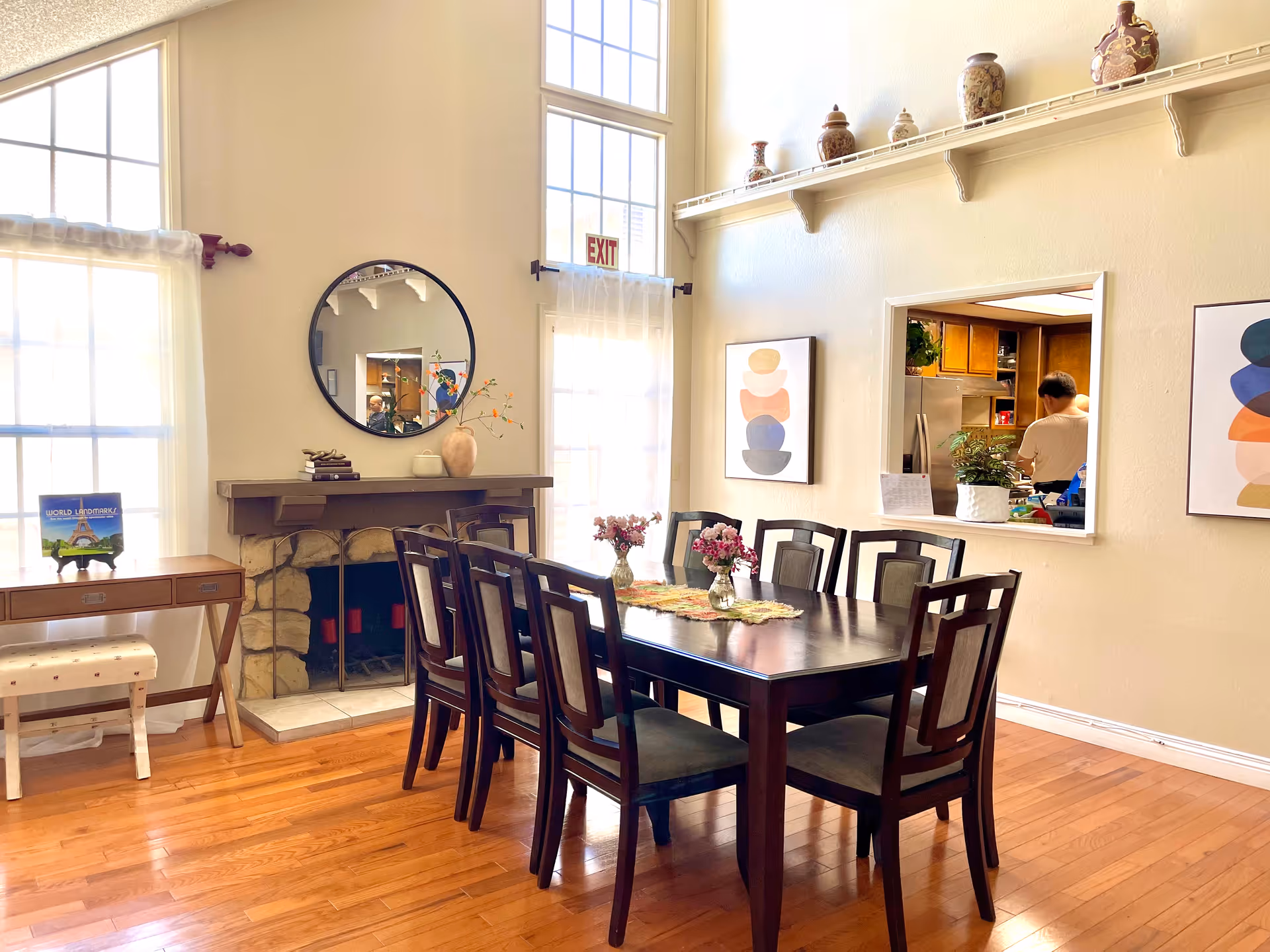A bright dining room with a dark wooden table and eight matching chairs. The room features large windows with sheer curtains, a stone fireplace with a round mirror above it, and decorative vases on a high shelf. There are two abstract art pieces on the wall and a pass-through window showing a person in the kitchen area.
