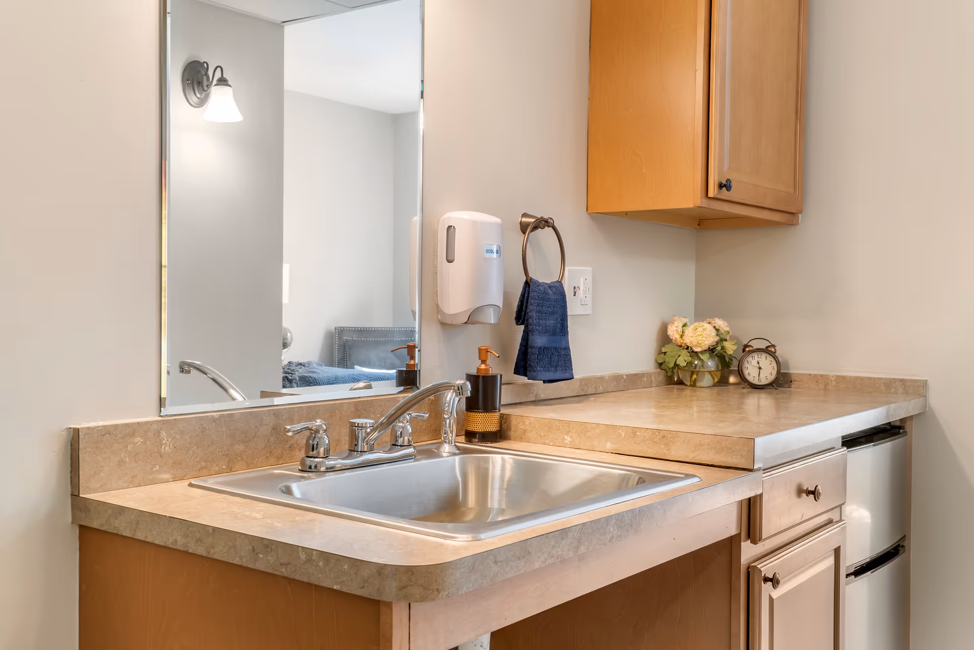 A clean bathroom sink area with a stainless steel faucet and sink, beige countertop, wooden cabinets, a wall-mounted soap dispenser, a towel ring with a blue hand towel, a small vase with flowers, and a vintage-style alarm clock. A large mirror reflects part of a bed and a wall lamp in the adjacent room.
