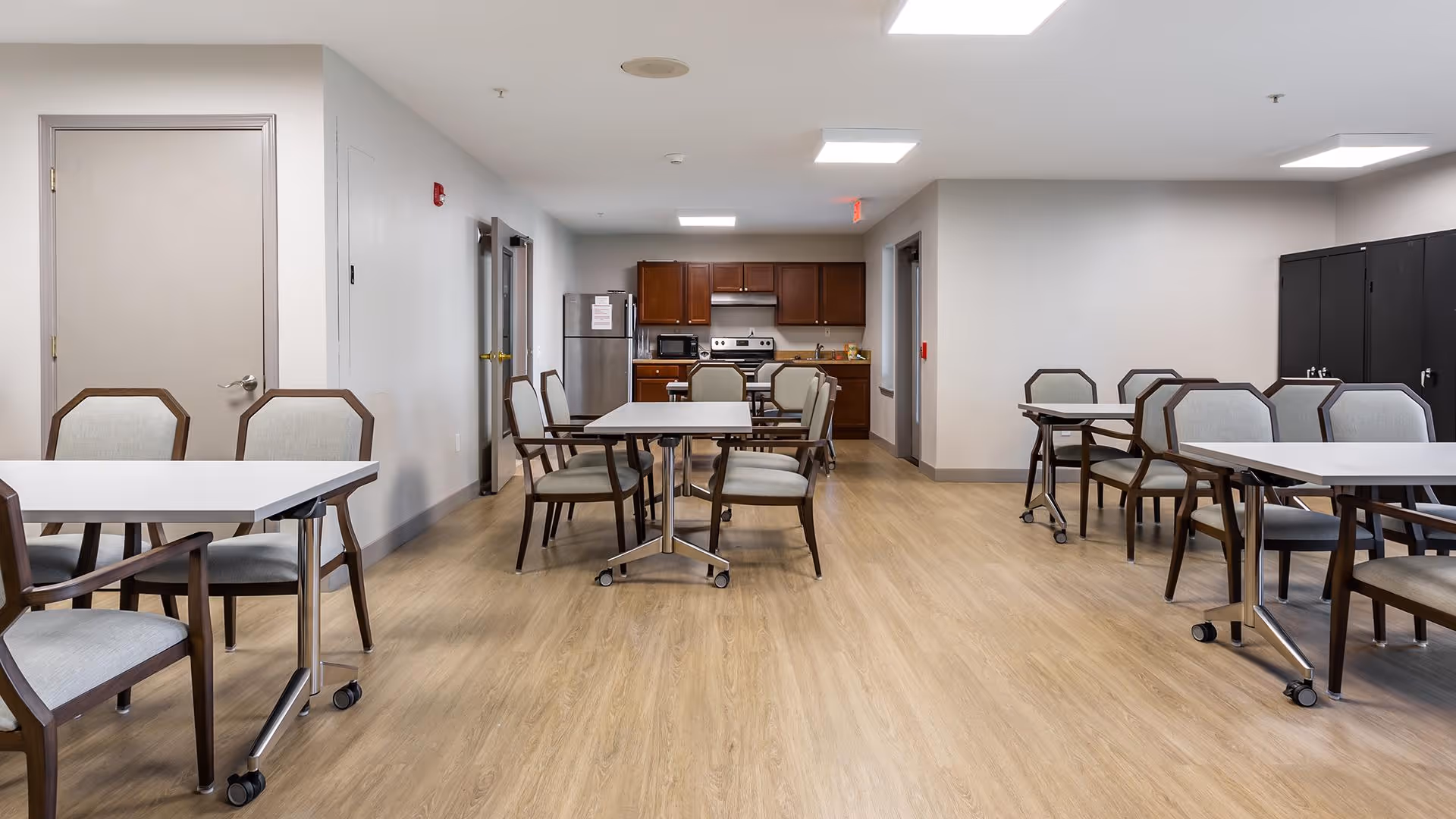 A spacious common dining area in a senior living facility with several tables and chairs arranged neatly. The room has light wood flooring, white walls, and a kitchen area with wooden cabinets, a refrigerator, microwave, and stove in the background. The ceiling has recessed lighting, and there are doors along the walls.