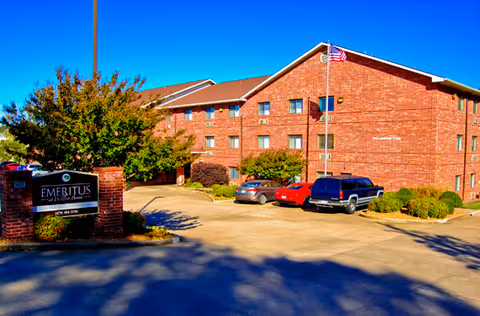 Exterior view of a three-story red-brick senior living building with parked cars, an 'Emeritus' entrance sign, and an American flag.