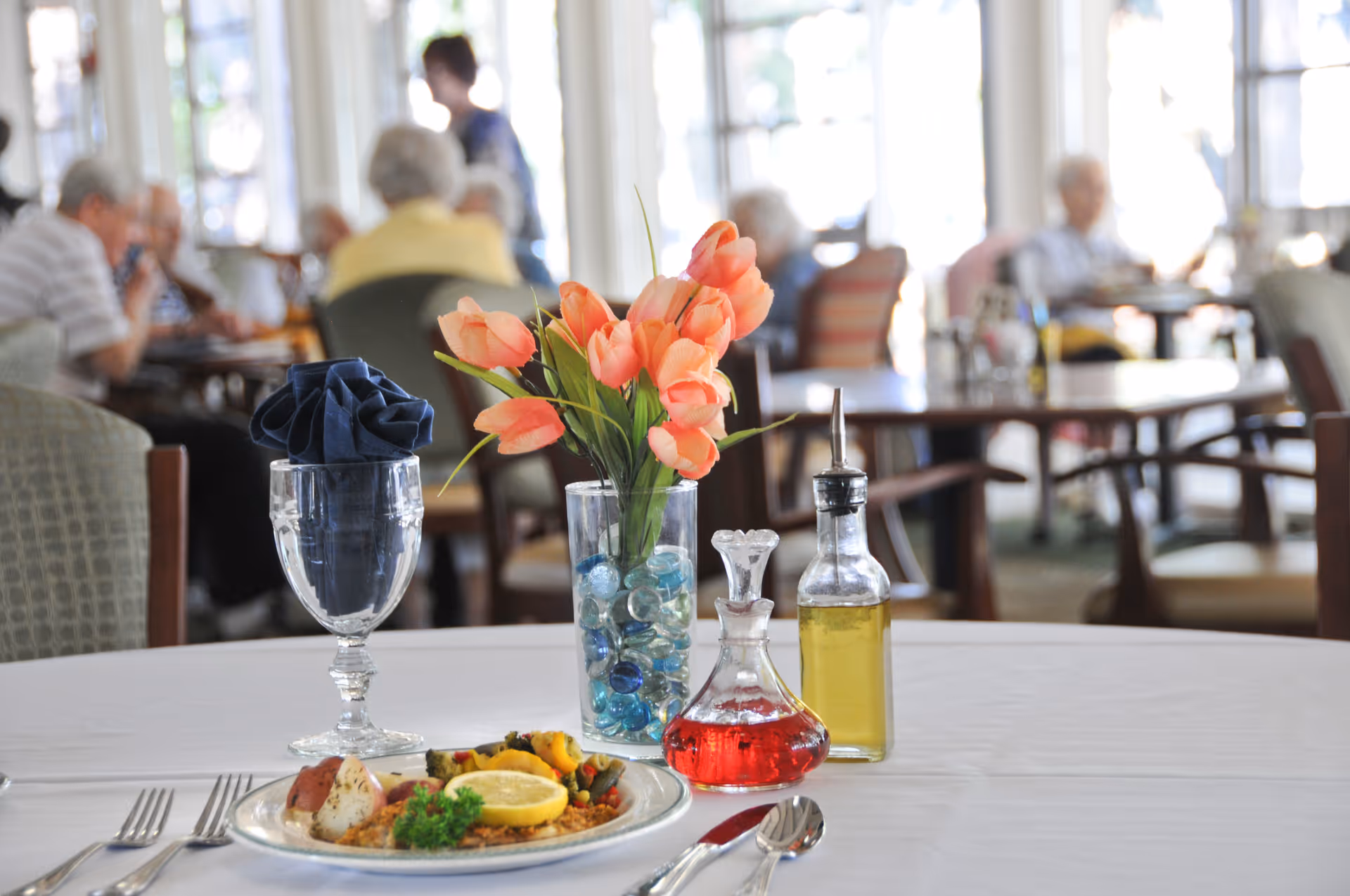 A dining table set with a plate of food, a glass with a folded navy blue napkin, a vase with orange tulips and decorative blue stones, and bottles of red and yellow condiments. In the background, elderly people are seated and dining in a bright room with large windows.