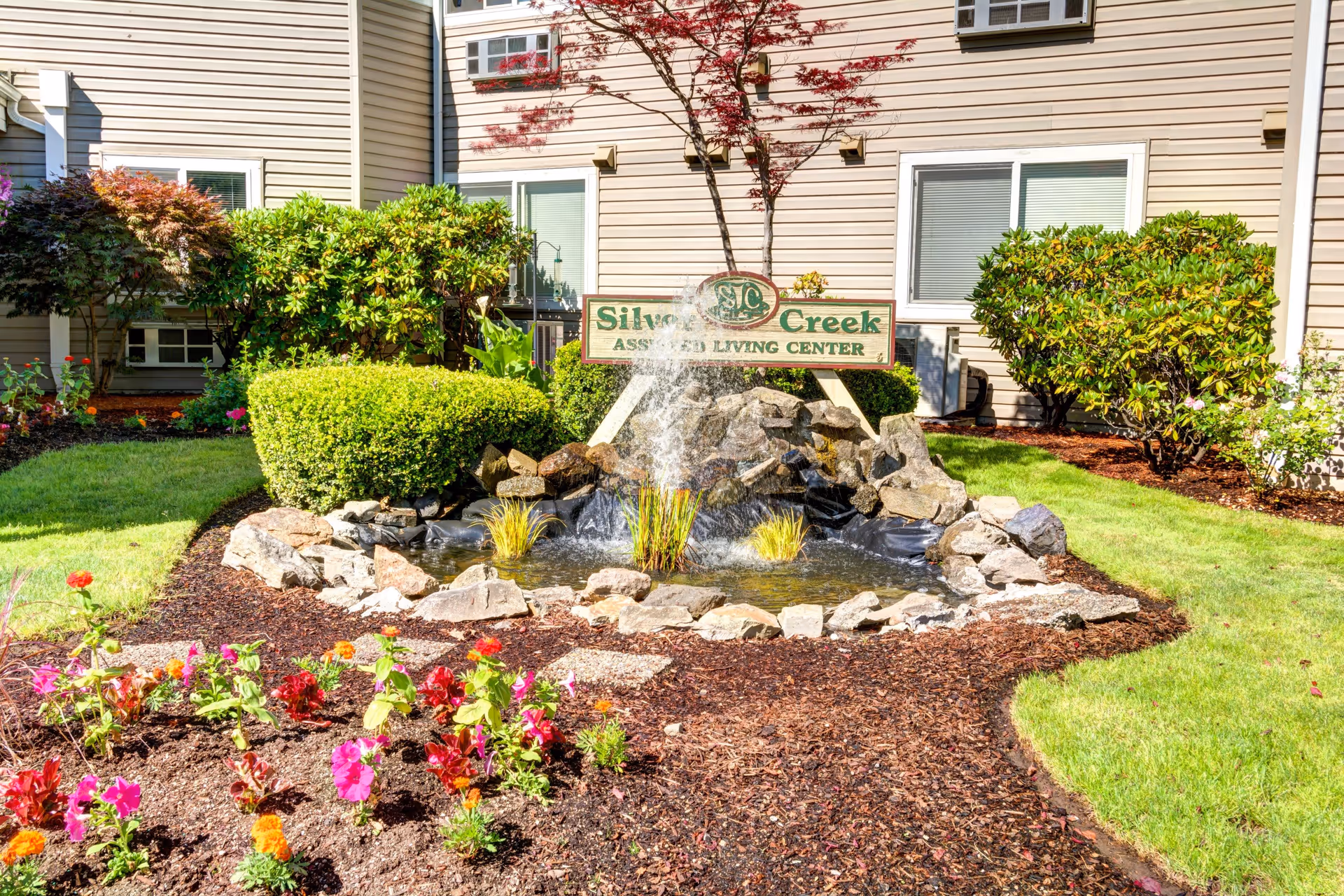 A landscaped garden area in front of a beige building with windows. The garden features a small pond with a water fountain surrounded by rocks, green bushes, and colorful flowers. A sign above the pond reads 'Silver Creek Assisted Living Center.'