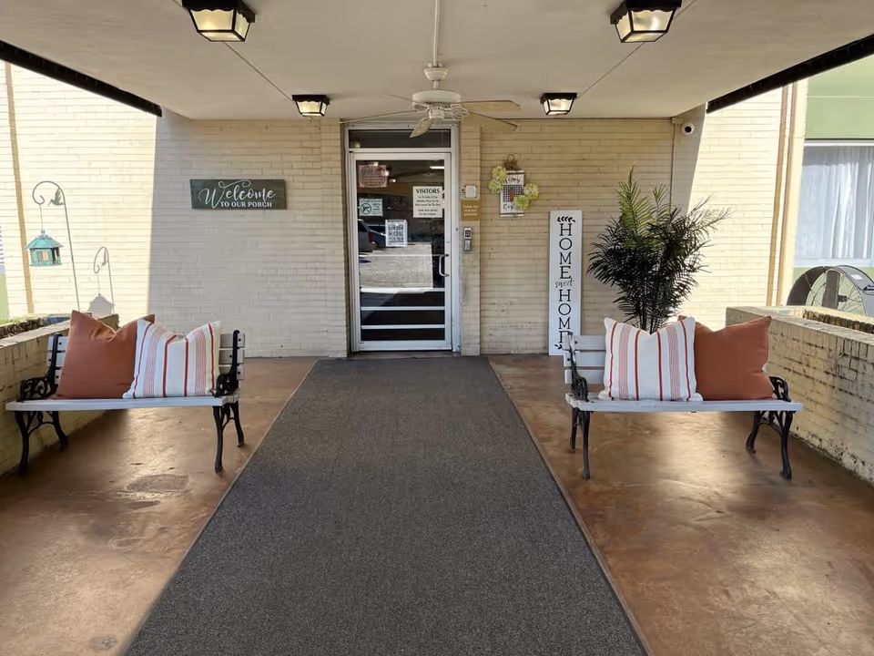 Covered porch entrance of a facility with two benches on either side, each with two pillows. A welcome sign is on the left wall, and a tall 'Home Sweet Home' sign is on the right next to a potted plant. The entrance door is glass with various notices posted on it, and there are ceiling lights and a ceiling fan above.