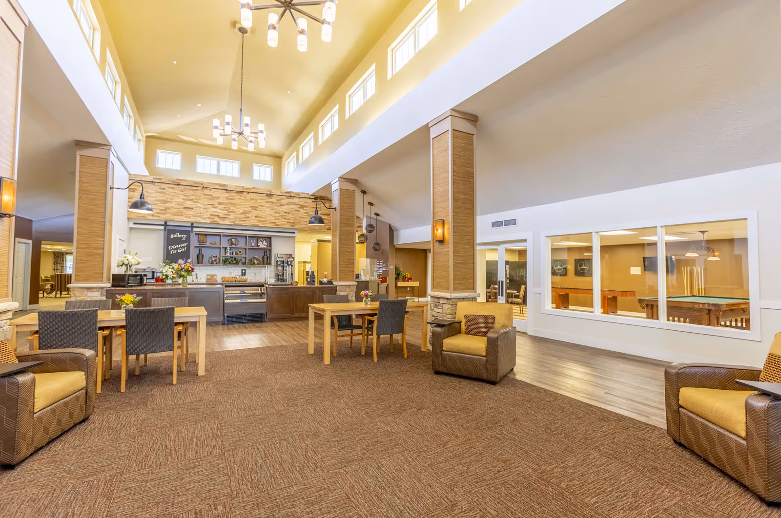 Bright, open communal dining and lounge area with high ceilings, tables, chairs, a serving counter, and seating.