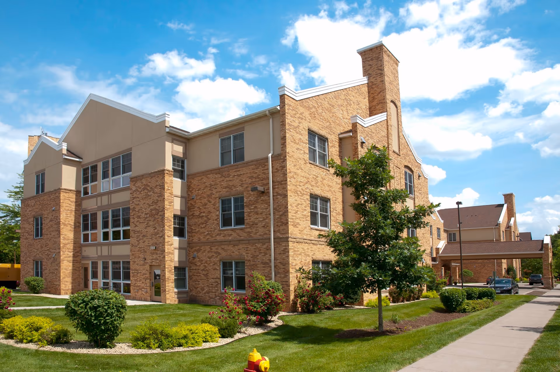 Exterior view of a multi-story brick building with large windows, surrounded by well-maintained green lawns, bushes, and a tree under a partly cloudy blue sky. A sidewalk runs alongside the building, and a covered entrance with parked cars is visible in the background.