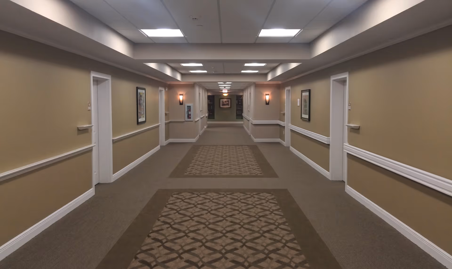 A long, well-lit hallway in a senior living facility with beige walls, white trim, and patterned carpet runners. Several closed white doors line both sides of the hallway, with framed artwork and wall sconces providing decoration and lighting.