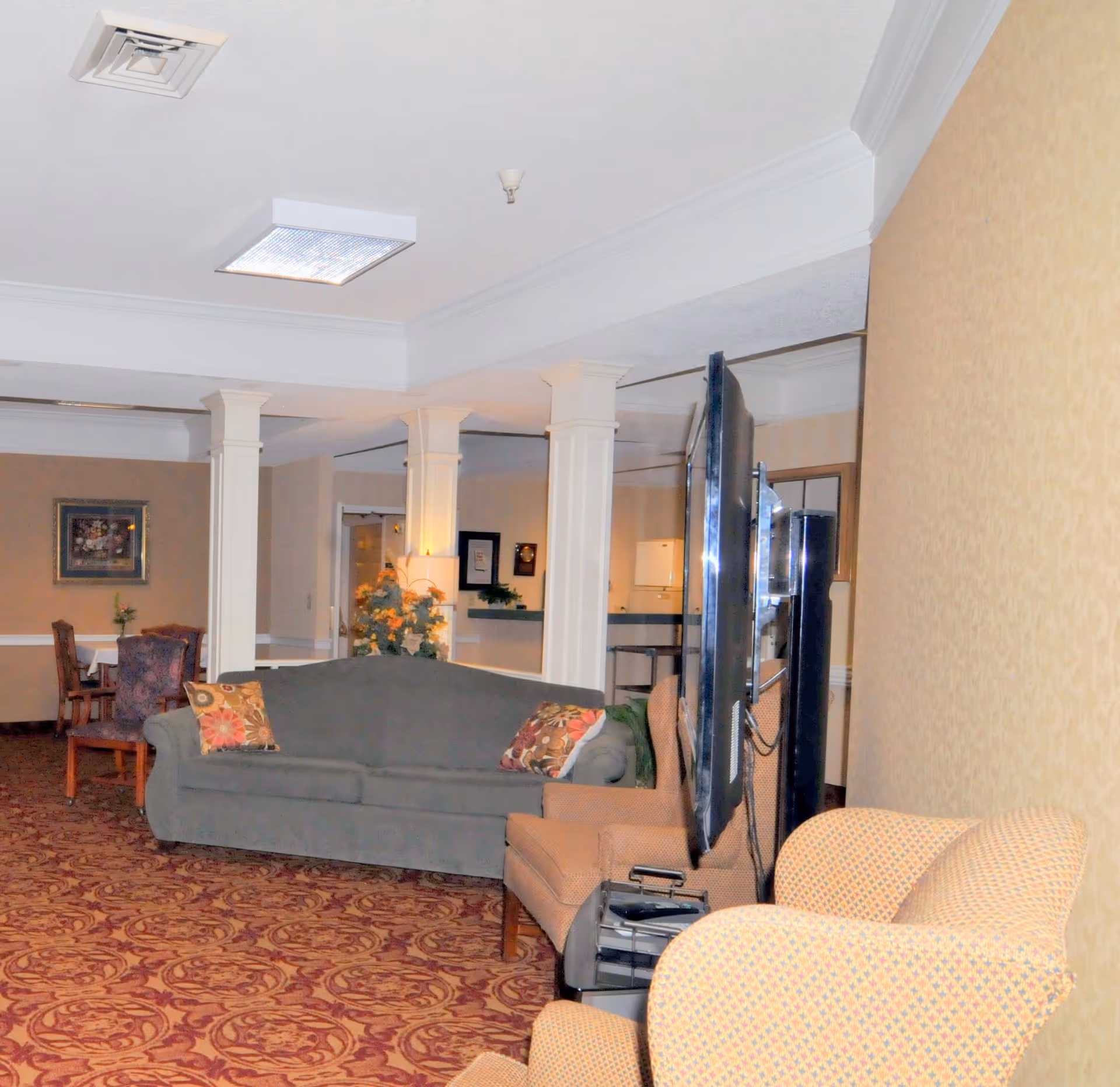 Interior view of a senior living facility lounge area with a gray sofa adorned with patterned pillows, two beige armchairs, a large flat-screen TV, and a carpet with an intricate red and gold pattern. In the background, there are white columns, a small dining table with chairs, framed artwork on the wall, and a floral arrangement near the columns.