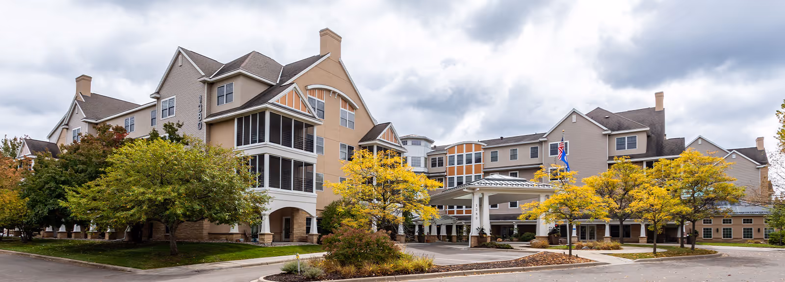 Exterior view of The Commons on Marice senior living facility showing a large multi-story building with beige and tan siding, multiple windows, and a covered entrance. There are trees with green and yellow leaves in front of the building and a cloudy sky overhead.