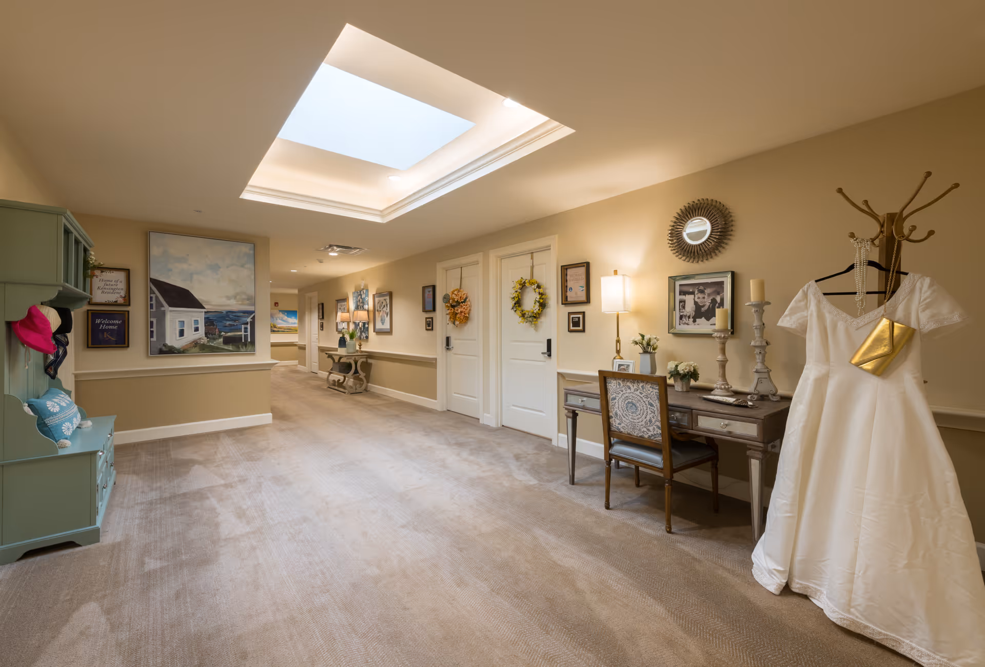 A well-lit hallway in a senior living facility with beige walls and carpeted floor. The hallway features a skylight, framed artwork, and decorative wreaths on two white doors. On the right side, there is a wooden desk with a chair, candles, flowers, and a framed photo. A white dress with a gold purse hangs on a coat rack nearby. On the left side, there is a green bench with pillows and hats.