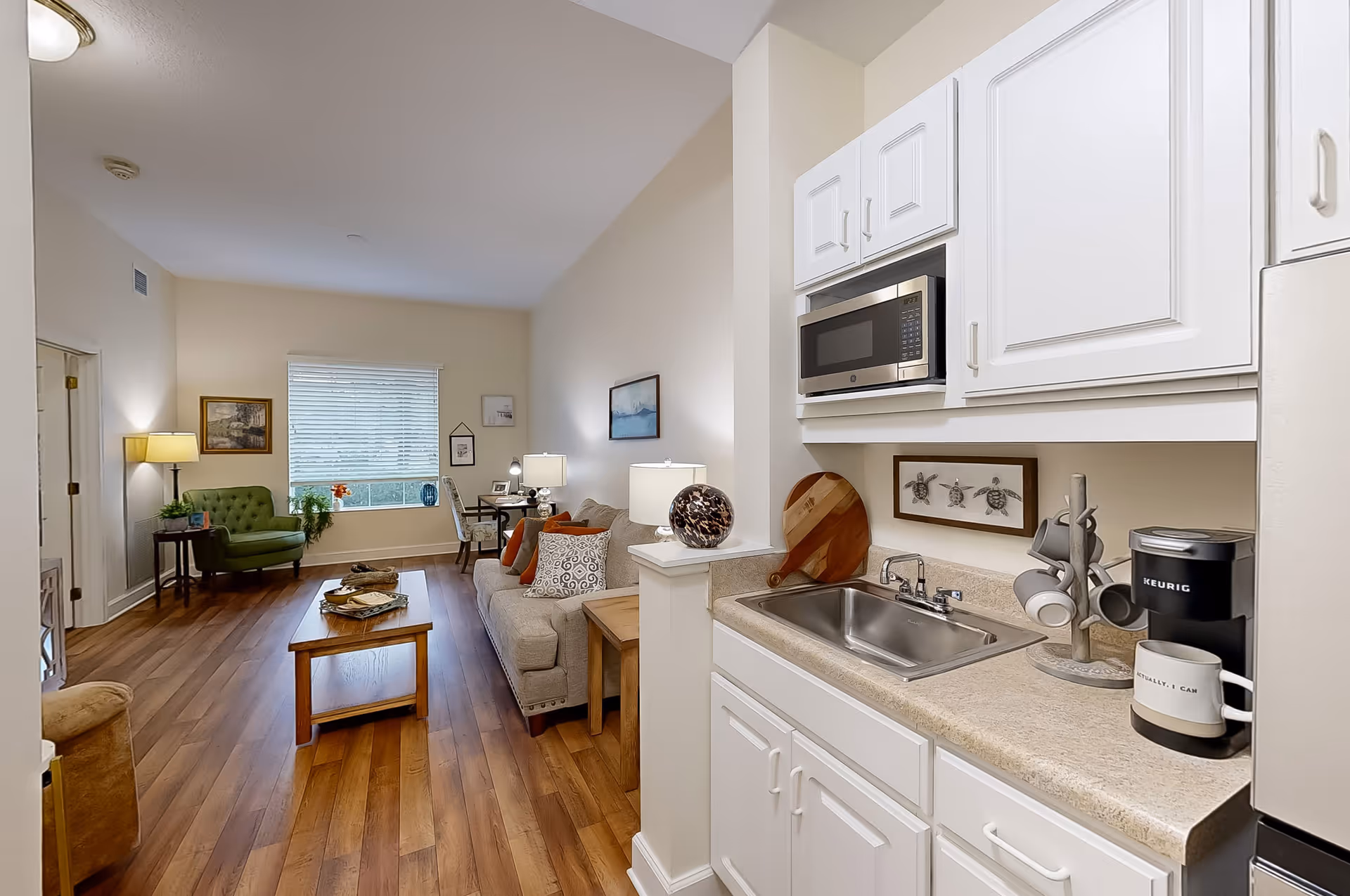 A bright and cozy living area with wooden flooring, featuring a beige sofa with patterned cushions, a wooden coffee table, a green armchair near a window with blinds, and a small desk with a chair. To the right is a compact kitchen area with white cabinets, a microwave, a sink, a Keurig coffee maker, and a mug on the counter. The walls are decorated with framed artwork.