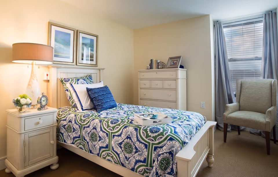 Sunlit bedroom with a patterned bed, nightstand with a lamp and clock, a dresser, and an armchair by a window.