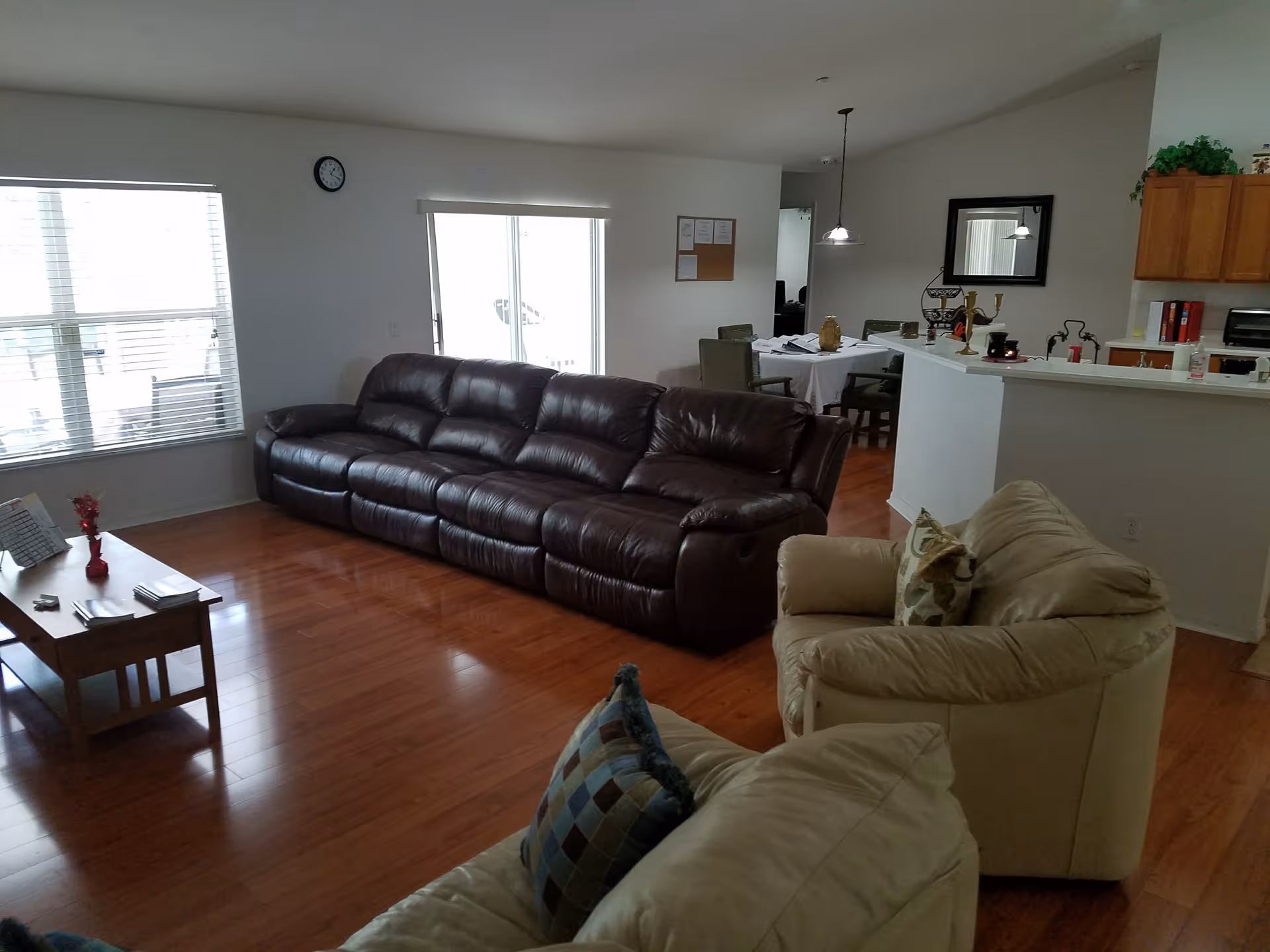 A living room area with a long dark brown leather sofa and two beige armchairs with patterned cushions. The room has wooden flooring and white walls. There is a wooden coffee table with some items on it near a window with blinds. In the background, a dining area with a table covered with a white tablecloth and green chairs is visible, along with a kitchen counter and cabinets. A clock is mounted on the wall above the sofa.