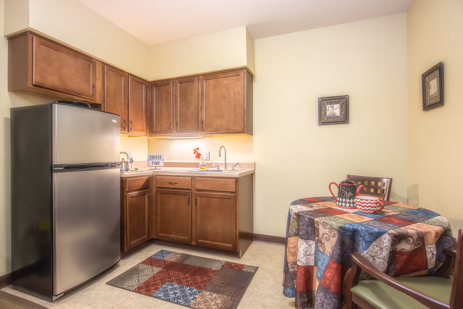 A small kitchen area with wooden cabinets, a stainless steel refrigerator, a sink, and a countertop. There is a colorful patchwork tablecloth on a round table with two chairs, a teapot, and a cup. The walls are light yellow with two framed pictures hanging.