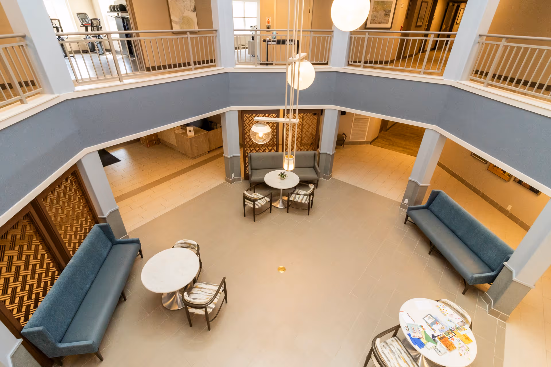 View from above of a spacious, modern common area in a senior living facility with blue cushioned benches, round tables with chairs, hanging globe lights, and a second-floor balcony overlooking the space.