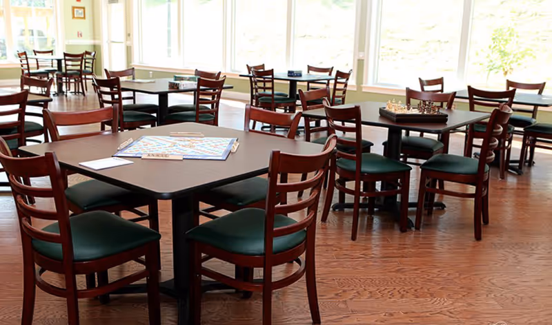 A bright dining room with multiple wooden tables and chairs arranged neatly. Some tables have board games like Scrabble and chess set up on them. Large windows let in natural light, illuminating the wooden floor and green cushioned chairs.