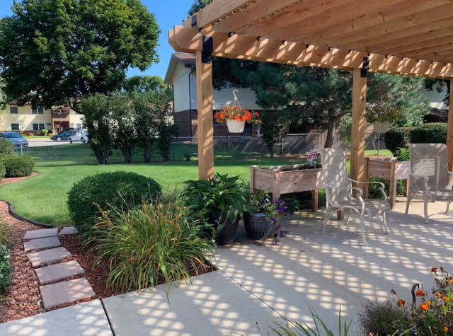 Sunlit outdoor patio with a wooden pergola, potted plants and chairs overlooking a landscaped lawn and neighboring building.