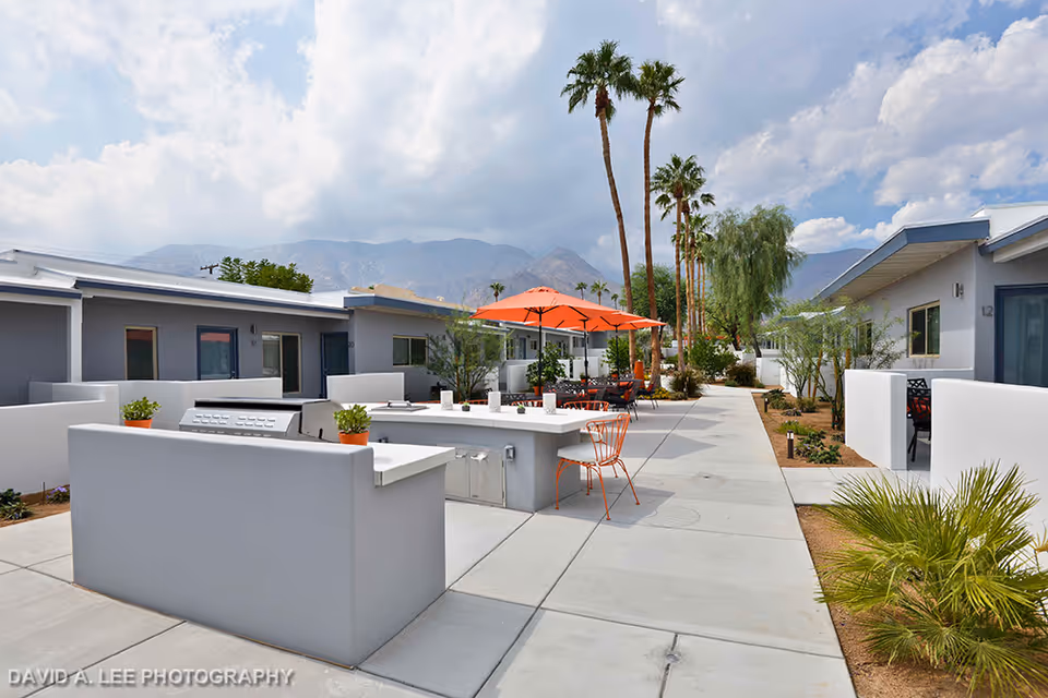 Outdoor courtyard area at Stonewall Gardens Assisted Living featuring a concrete walkway, modern white low walls, a built-in barbecue grill, orange patio umbrellas, metal chairs, and palm trees with mountains in the background under a partly cloudy sky.