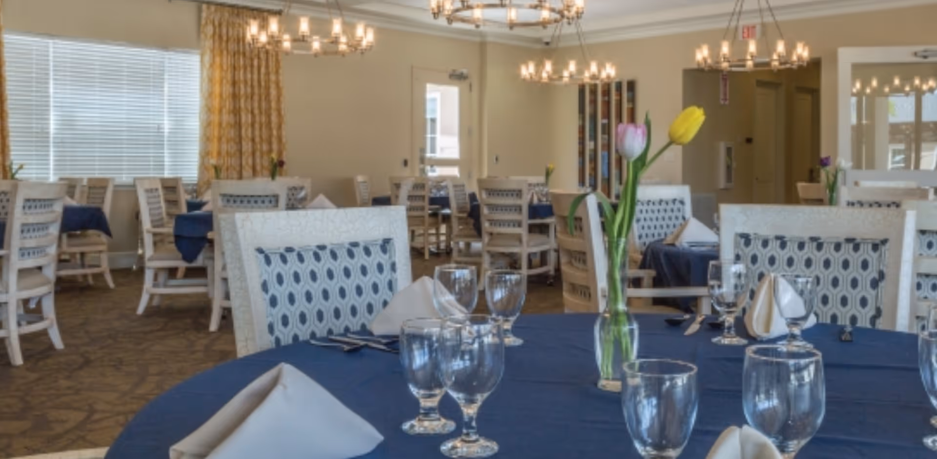 A dining room with multiple tables covered in dark blue tablecloths, each set with glassware, silverware, and folded napkins. The chairs have patterned upholstery, and there are chandeliers hanging from the ceiling. A vase with tulips is placed on the table in the foreground. Large windows with blinds and yellow curtains allow natural light into the room.