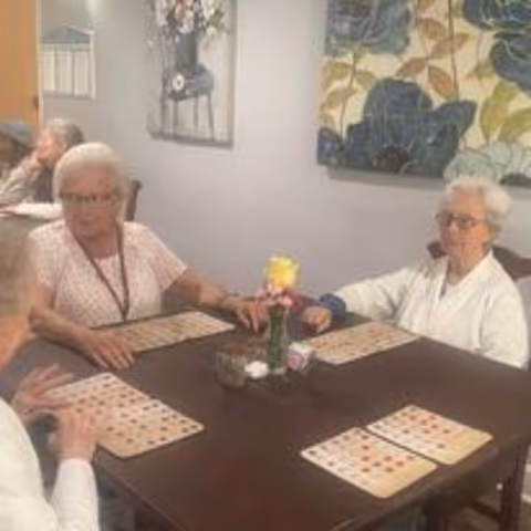 Four elderly women sitting around a dark wooden table playing bingo in a well-lit room with floral artwork on the walls.