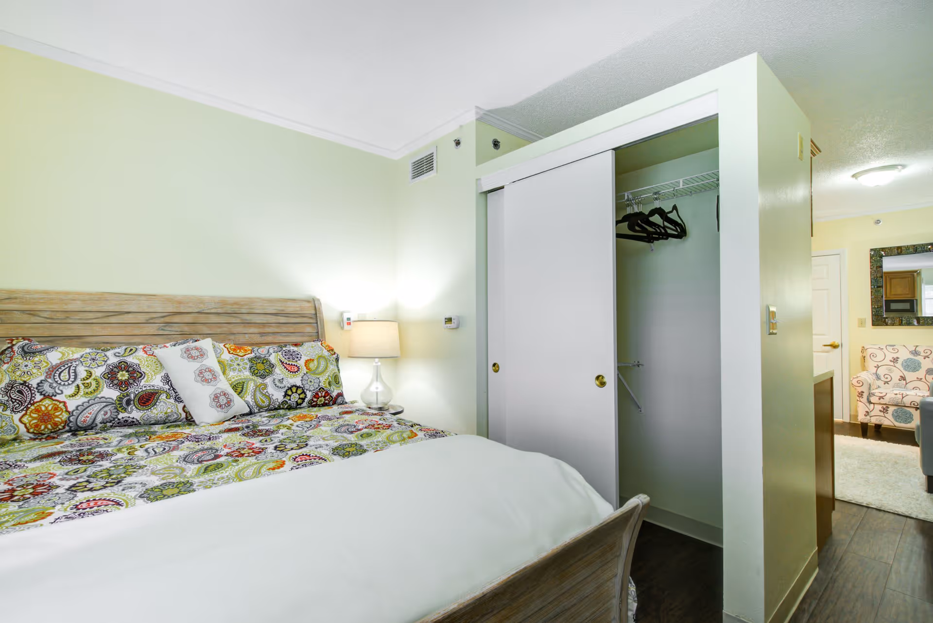 A bedroom with a wooden bed frame and colorful patterned bedding. There is a nightstand with a lamp next to the bed. A closet with sliding doors is partially open, showing hangers inside. In the background, there is a view into a living area with a patterned armchair, a mirror on the wall, and a ceiling light.