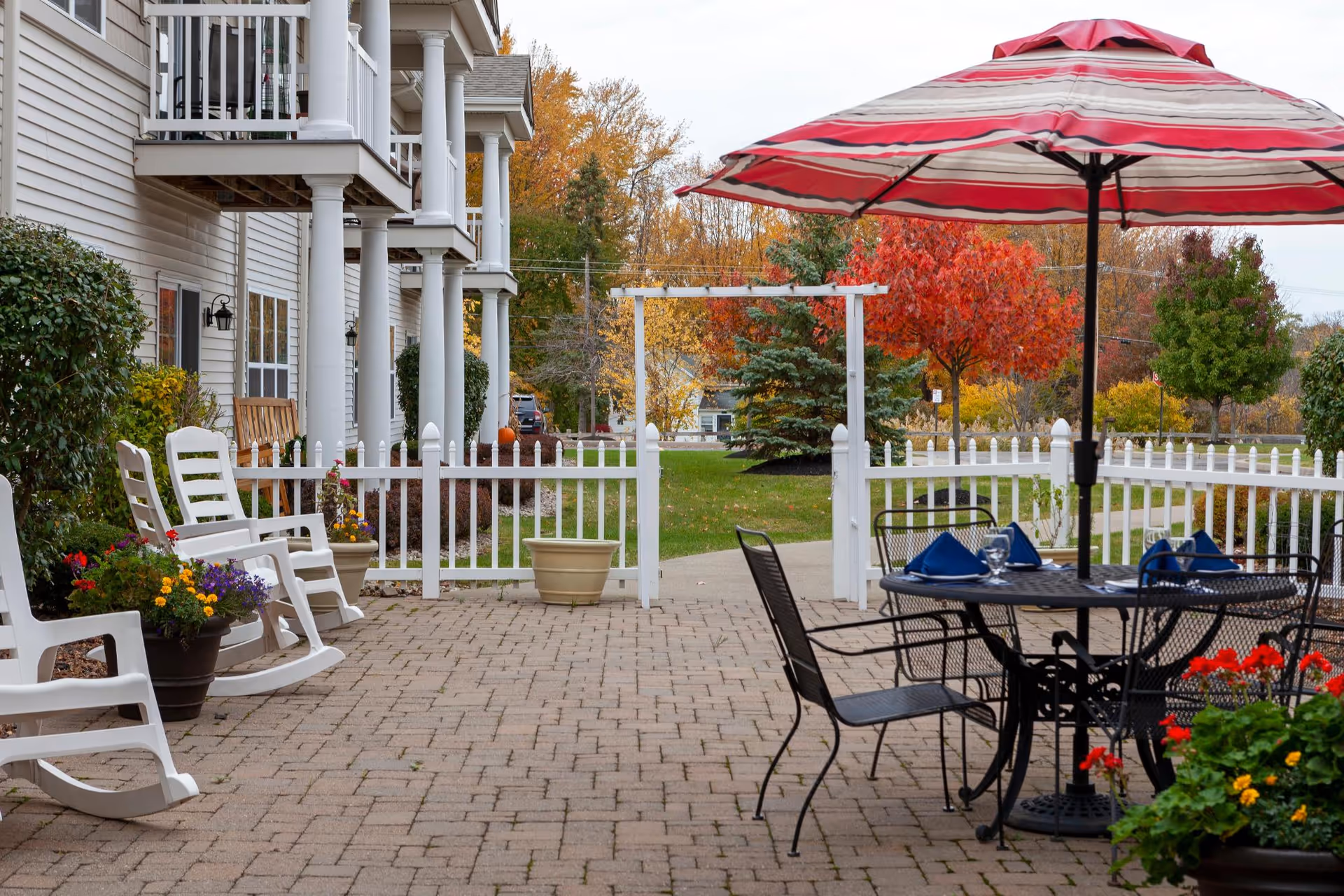 Patio with white rocking chairs, a table under a red-striped umbrella, potted flowers and a white picket fence beside a multi-story building with columns and autumn trees.