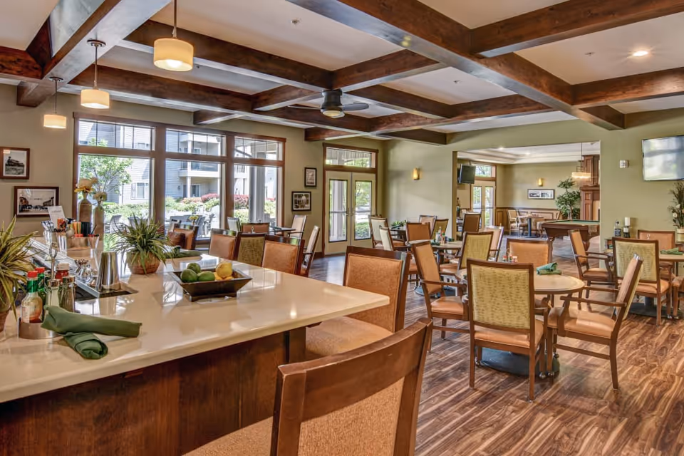 A spacious dining room in a senior living facility with multiple tables and chairs arranged neatly. The room features large windows allowing natural light to brighten the space, wooden ceiling beams, and a countertop with decorative plants and condiments. The flooring is wood-style, and there are ceiling fans and wall-mounted lights providing additional lighting.