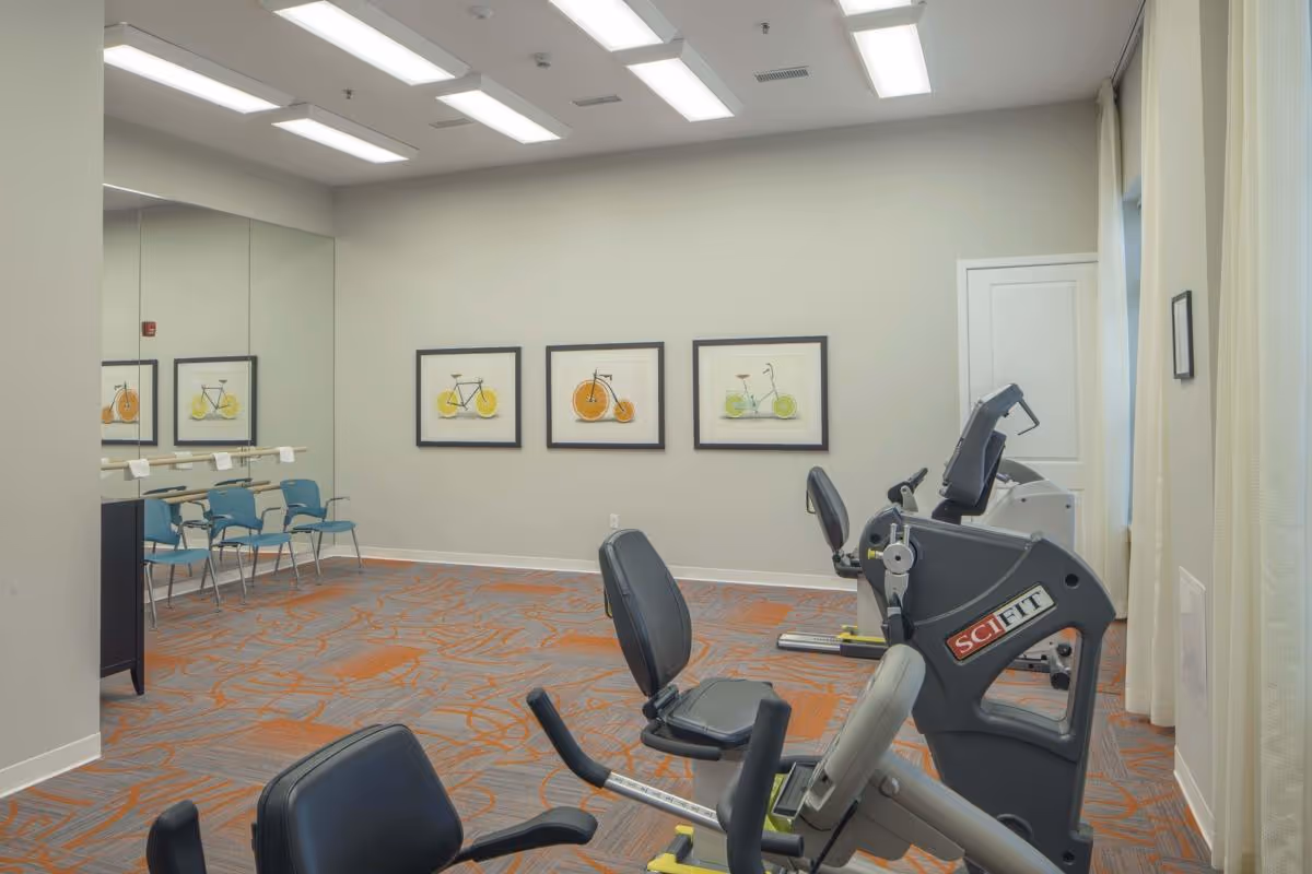 A small exercise room with two recumbent exercise bikes facing a wall with three framed bicycle-themed artworks. The room has a patterned carpet with orange and gray colors, a large mirror on one wall, and several blue chairs lined up against the mirrored wall. The ceiling has rectangular fluorescent lights, and there is a window with light-colored curtains on the right side.