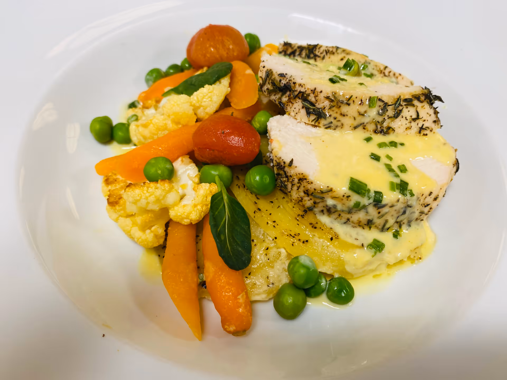 A plated meal featuring two slices of herb-crusted chicken breast topped with a creamy yellow sauce and chopped chives, served with a side of cooked baby carrots, peas, cauliflower, cherry tomatoes, and a grilled yellow vegetable on a white plate.