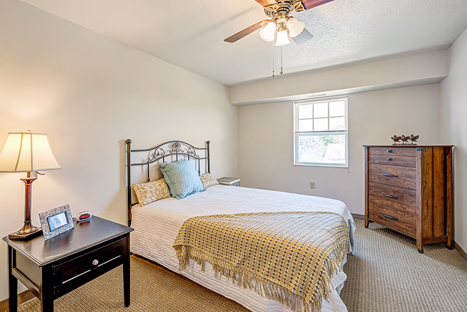 Bright furnished bedroom with a bed, nightstand and lamp, a chest of drawers, and a window.