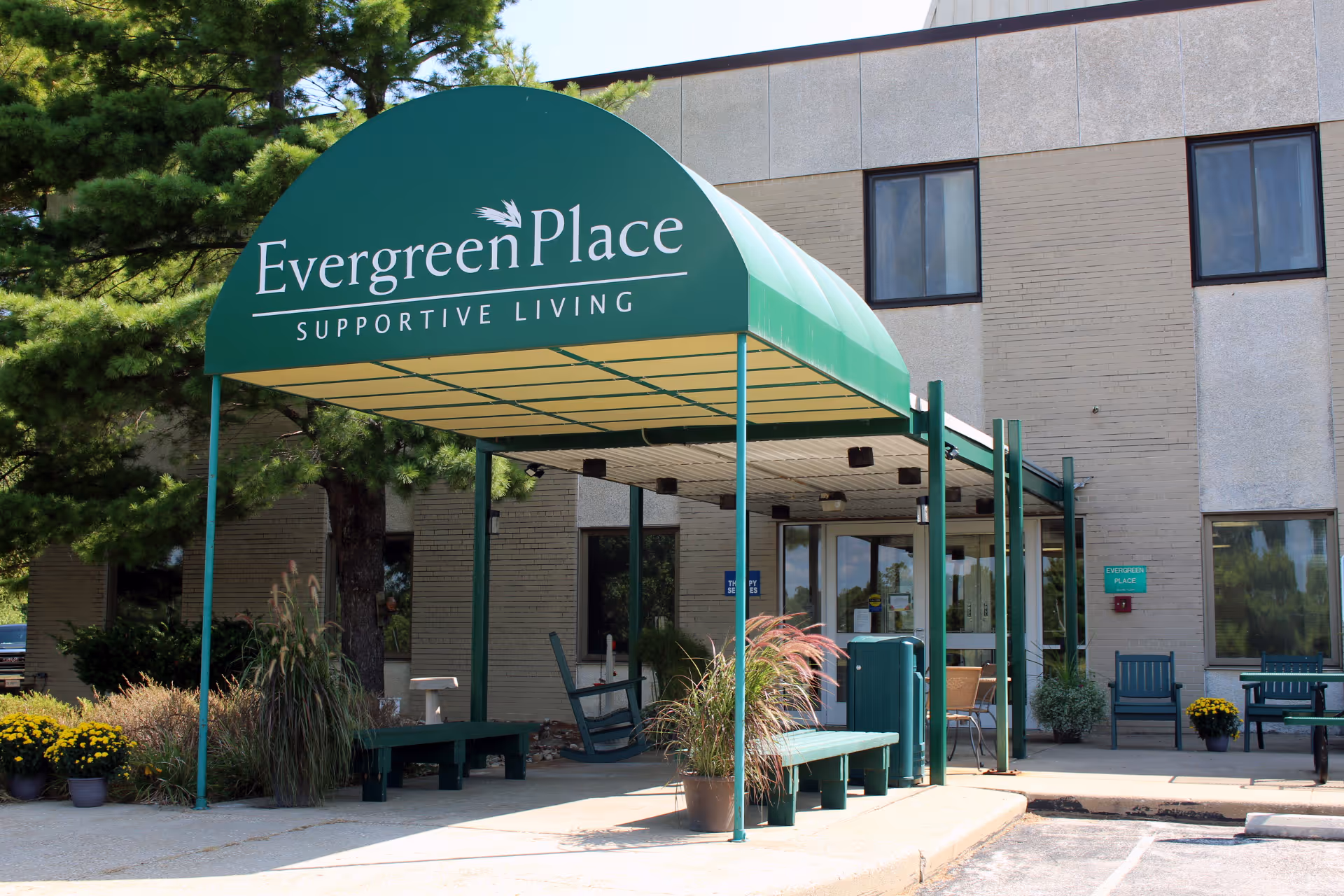 Entrance to Evergreen Place Supportive Living facility with a green canopy over the walkway, benches, potted plants, and a beige brick building in the background.