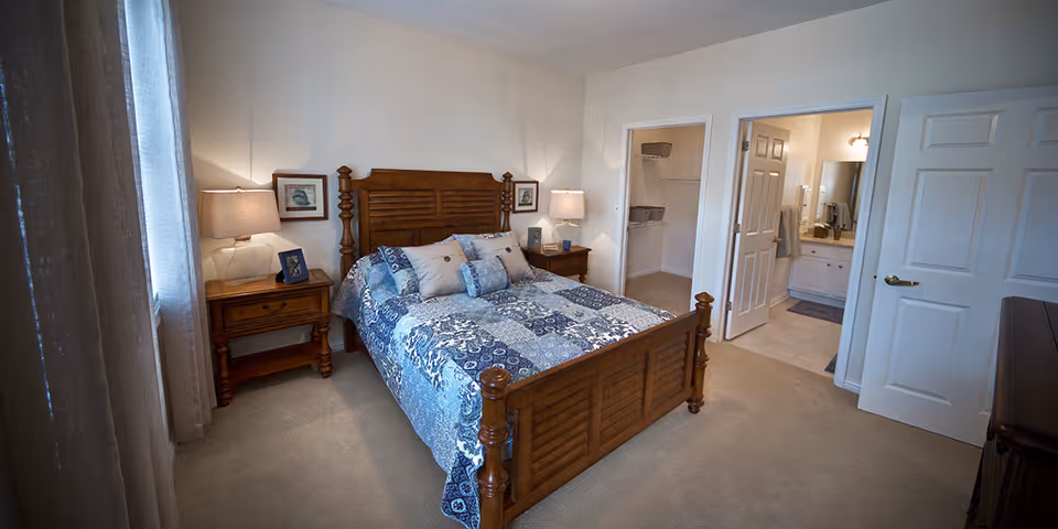 A bedroom with a wooden bed frame and a blue patterned quilt. There are two wooden nightstands with lamps on either side of the bed. The room has beige carpet and light-colored walls. A walk-in closet and a bathroom with a sink and mirror are visible through open doors.
