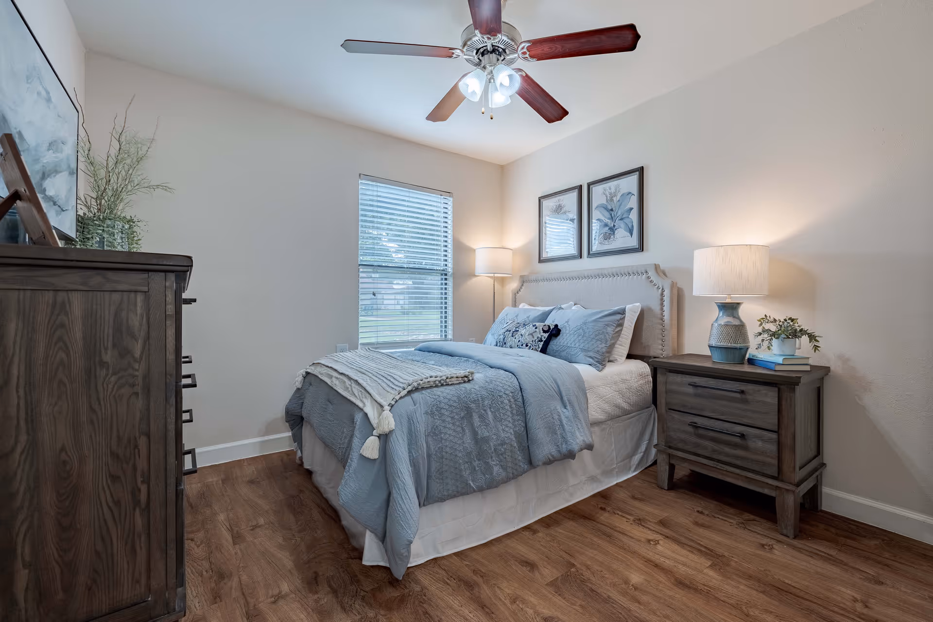 A neatly made bed with light blue bedding and decorative pillows in a bedroom. There is a wooden nightstand with a lamp, books, and a small plant on the right side of the bed. A wooden dresser with a plant and framed artwork is on the left side. The room has a window with blinds, light-colored walls, wood flooring, and a ceiling fan with lights.