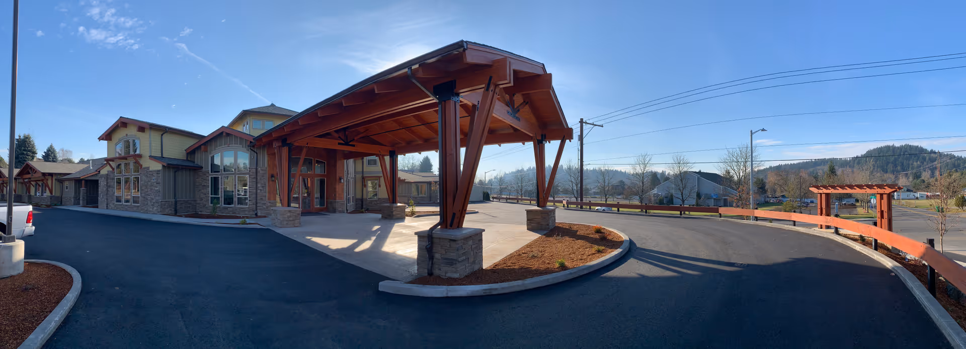 Wide panoramic view of the exterior entrance of Mountain Park Memory Care facility featuring a large covered drop-off area with wooden beams and stone pillars, a paved driveway, and surrounding landscaped areas with mulch. The background shows a clear blue sky and distant hills with trees.