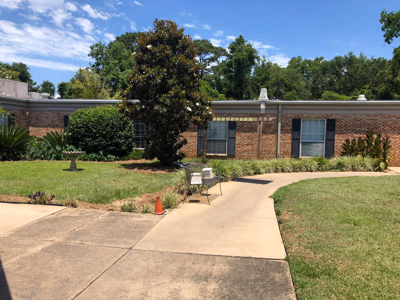 Outdoor view of Montrose Bay Health and Rehab showing a paved walkway curving through a grassy area with a bench and a small orange traffic cone. There are bushes, a birdbath, and a large tree near a single-story brick building with windows and black shutters under a partly cloudy blue sky.