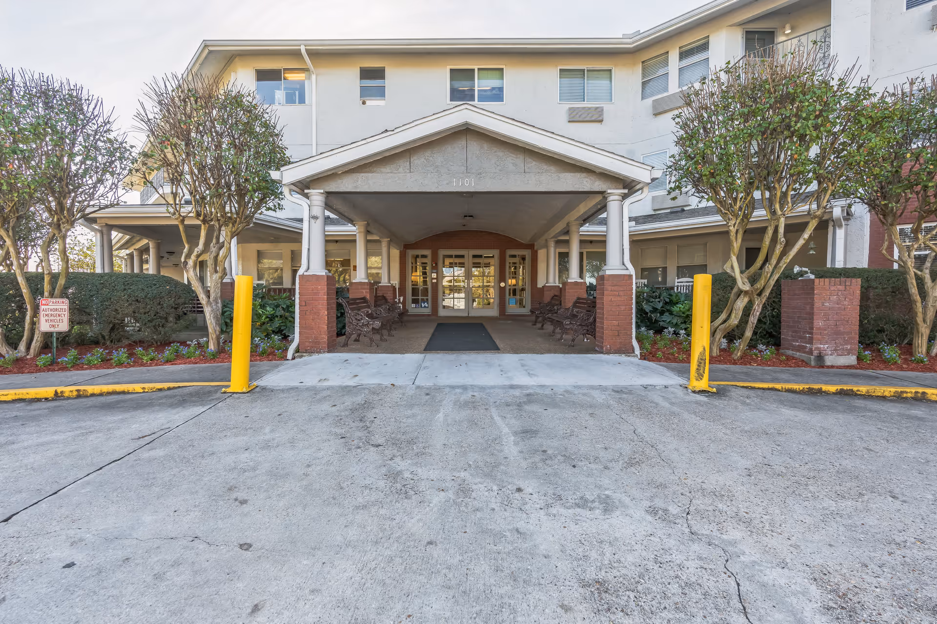 Front entrance of a senior living facility with a covered drop-off area supported by white columns and brick bases. There are benches on either side under the covered area, small trees and bushes lining the walkway, and a sign indicating no parking except for authorized emergency vehicles.