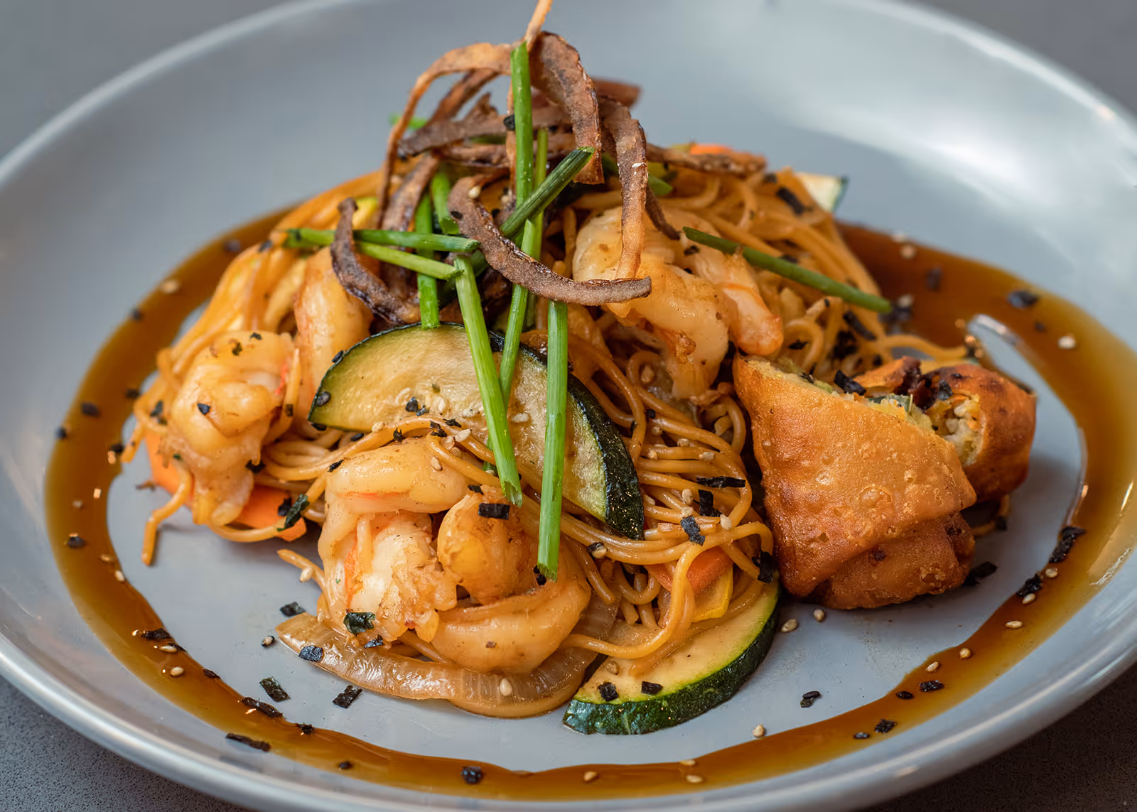 A plate of Asian-inspired food featuring shrimp, noodles, sliced zucchini, and garnished with green chives and crispy fried strips, served with a dipping sauce on the side.