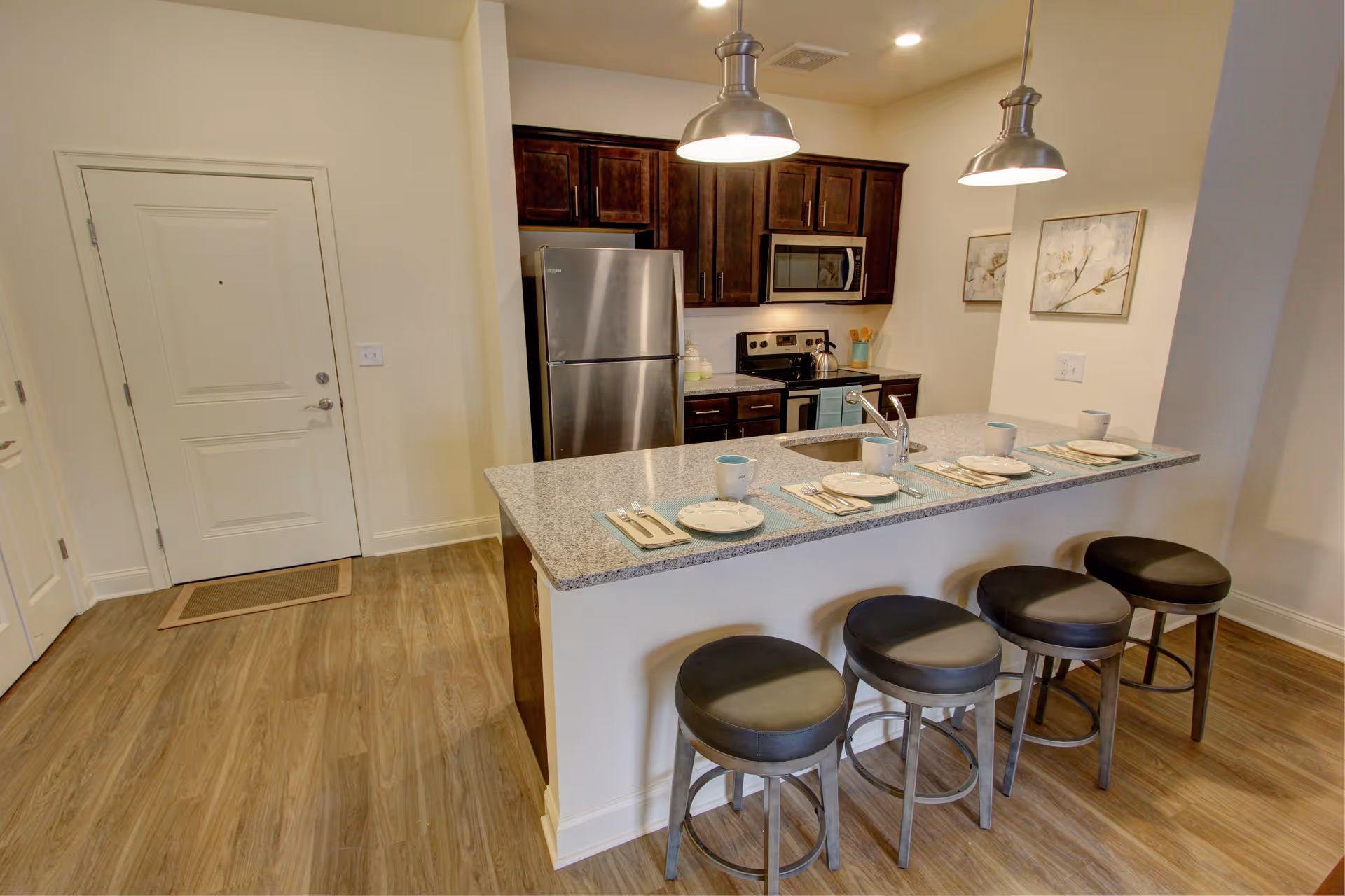 Modern kitchen area with a granite countertop island featuring four black cushioned stools. The countertop is set with four place settings including plates, cups, and cutlery. The kitchen has dark wood cabinets, a stainless steel refrigerator, microwave, and stove. The floor is wood, and the walls are light-colored with two framed artworks. There are two pendant lights hanging above the island.