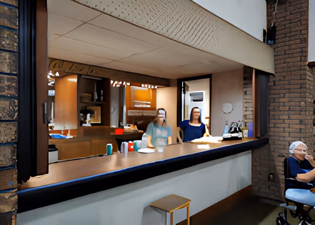 Interior view of a senior living facility kitchen area with two women standing behind a counter. The counter has various items including cups and plates. To the right, an elderly person is seated in a wheelchair near a brick wall.