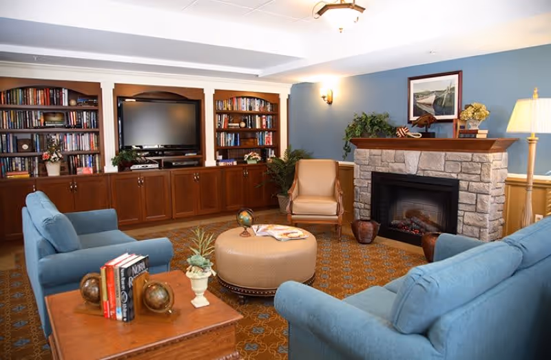 A cozy living room with blue upholstered sofas and a beige armchair arranged around a round ottoman. There is a wooden coffee table with books and decorative globes. The room features a stone fireplace with a wooden mantle, a framed picture above it, and a floor lamp. Built-in wooden bookshelves filled with books and a flat-screen TV are on the back wall. The walls are painted blue with white trim and the floor is covered with a patterned carpet.