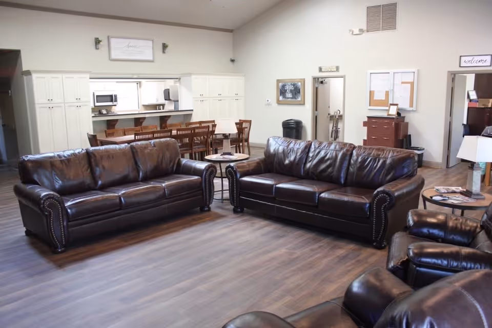 Spacious common living area with two brown leather sofas and armchairs facing a seating area, with a dining table and kitchenette visible in the background.