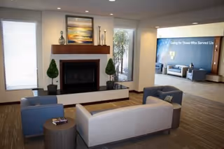 A modern assisted living facility common area with a fireplace centered between two windows. There are two small potted topiary plants on either side of the fireplace. In front of the fireplace, there is a seating arrangement consisting of a gray sofa, two blue armchairs, and a small round wooden table with magazines. In the background, another seating area with blue chairs and a blue wall with the text 'Caring for Those Who Served Us' is visible.