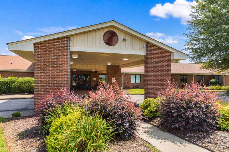 Front exterior view of a single-story brick building with a covered entrance supported by two brick columns. There are landscaped bushes and plants in front of the entrance, and a clear blue sky with some clouds above.