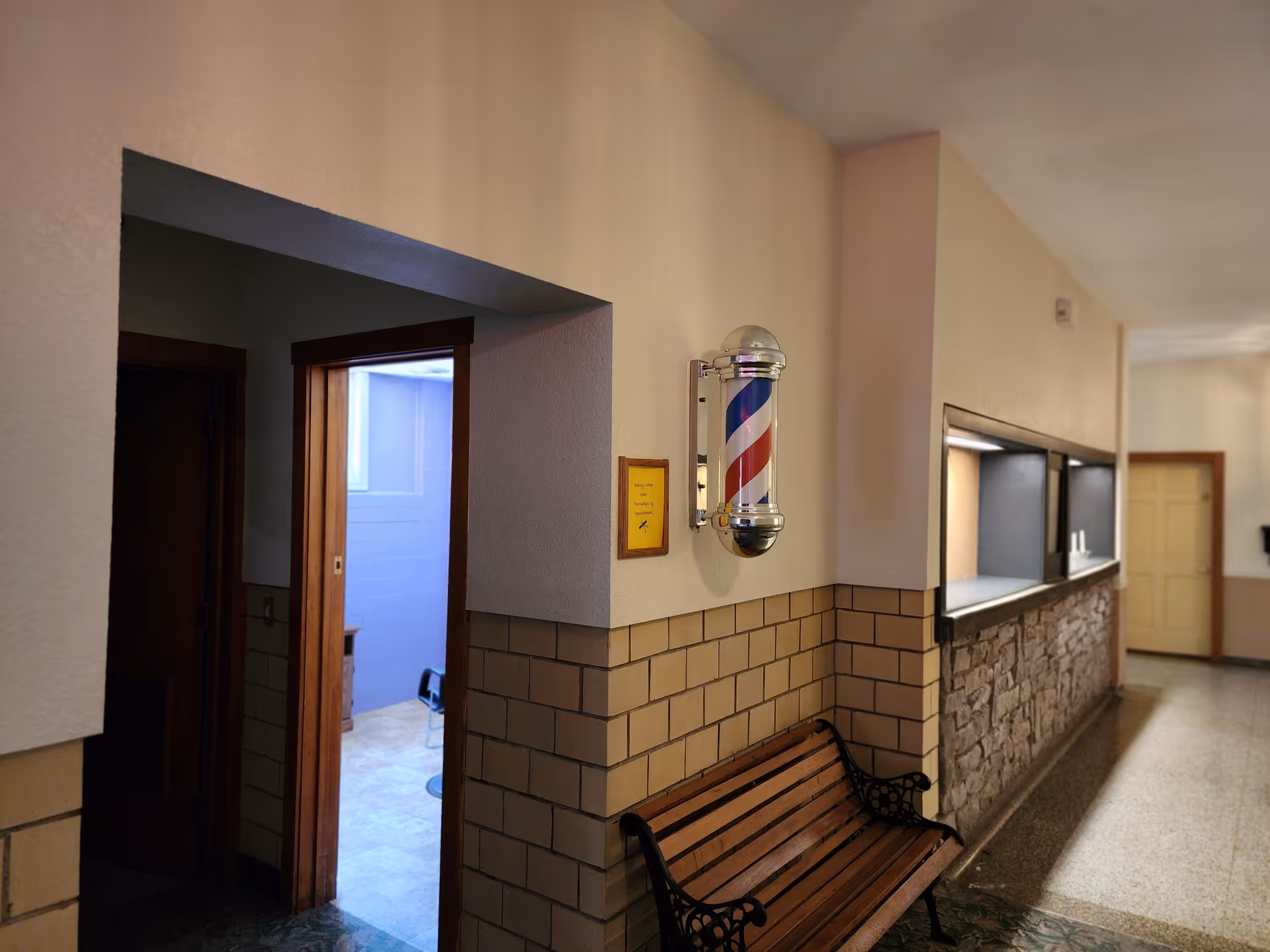 Indoor hallway with beige walls and tiled lower half, featuring a wooden bench with black metal armrests. A traditional barber pole is mounted on the wall above the bench. There is an open doorway leading to a room with light blue walls and a window. Further down the hallway, there is a counter with a stone facade and two open service windows. A closed beige door is visible at the end of the hallway.