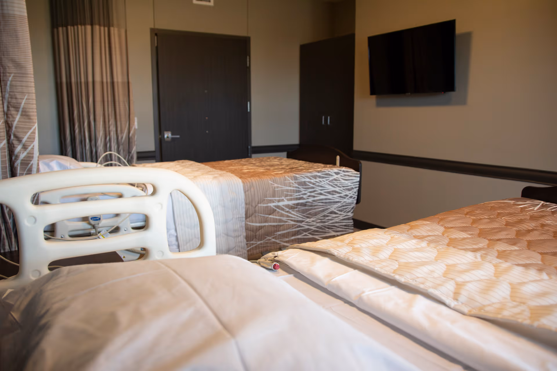 A shared senior living bedroom with two beds, a wall-mounted TV, and a closed door in the background.