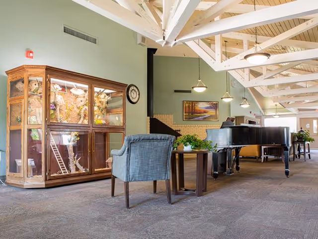 A spacious common area with a high ceiling featuring exposed white beams and hanging lights. The room includes a large black grand piano, a blue upholstered armchair, a wooden side table with a green plant, and a wooden display cabinet with glass doors containing decorative items. A clock is mounted on the wall above the cabinet, and a framed landscape painting hangs on the far wall near a fireplace.