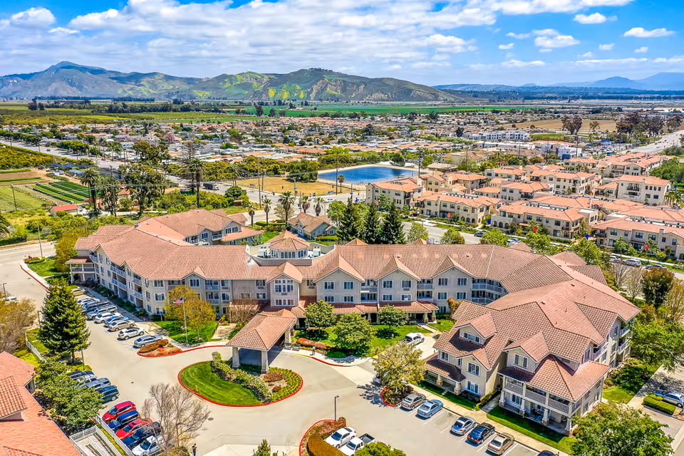 Aerial view of The Palms At Bonaventure Assisted Living & Memory Care facility showing a large, multi-wing building with red-tiled roofs surrounded by parking lots, green lawns, trees, and a circular driveway. In the background, there are residential neighborhoods, fields, and distant mountains under a partly cloudy sky.