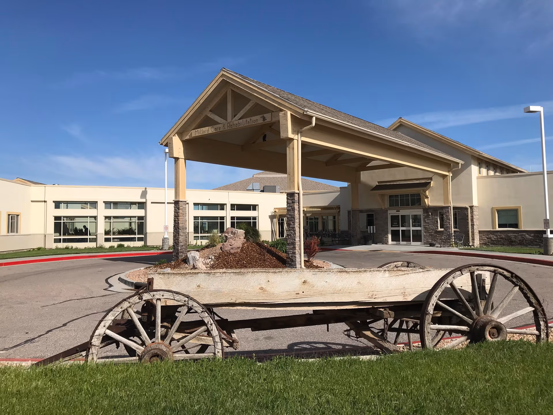Front exterior view of Millard County Care Center building with a covered entrance, stone pillars, and a wooden wagon displayed on the grass in the foreground under a clear blue sky.