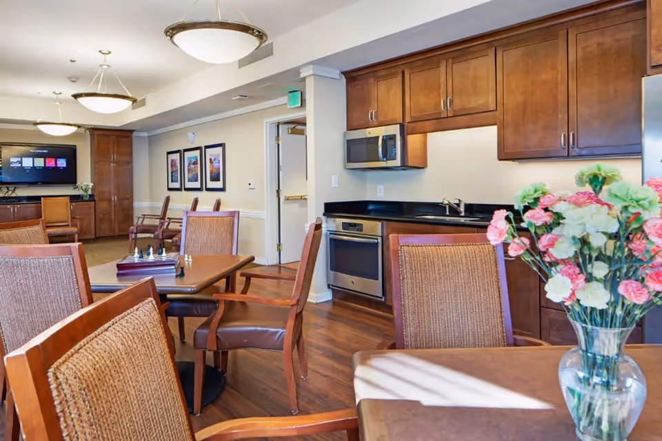 A communal dining and kitchen area with wooden tables and chairs, a chess set on one table, a vase with pink and white flowers on another table, wooden cabinetry, a microwave, an oven, and a flat-screen TV mounted on the wall in the background.