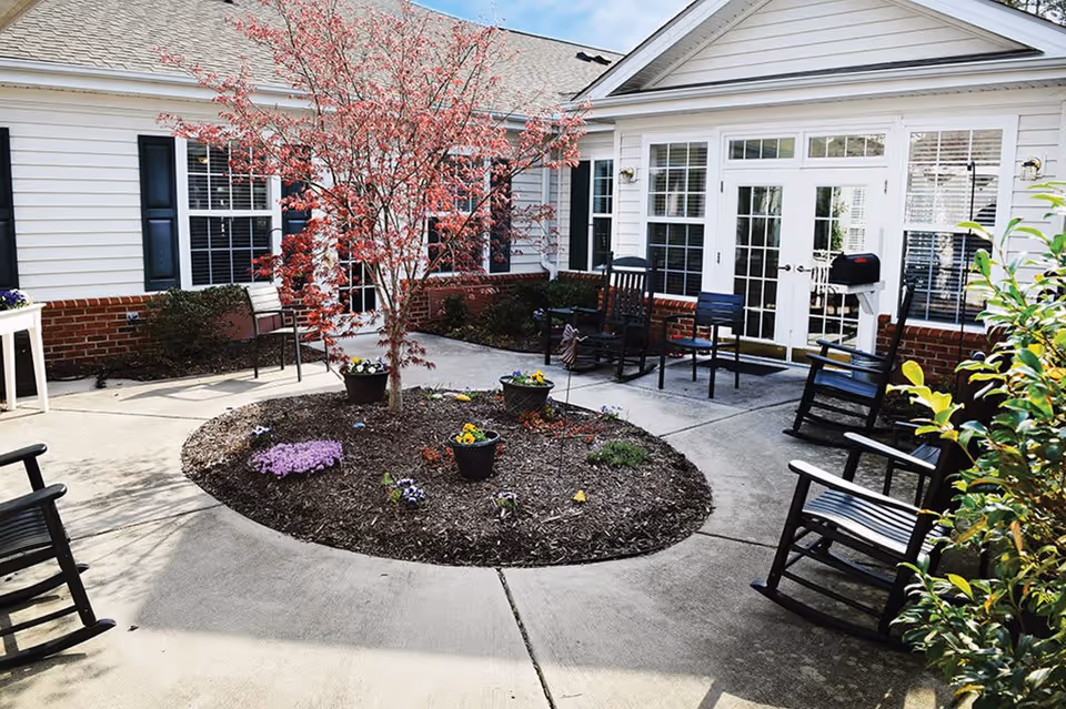 Outdoor courtyard area with a circular garden bed containing a small tree and several flower pots. Surrounding the garden bed are several black rocking chairs and a few regular chairs on a concrete patio. The background shows a white building with brick accents, multiple windows with black shutters, and glass double doors.